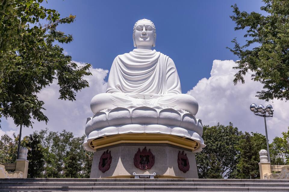 A large white statue of a seated figure with a lotus base, surrounded by trees and a clear sky.
