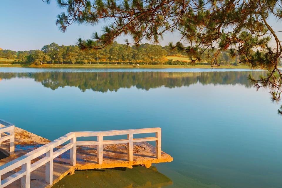 A calm lake with a wooden dock extending into the water, surrounded by trees and a distant mountain range.