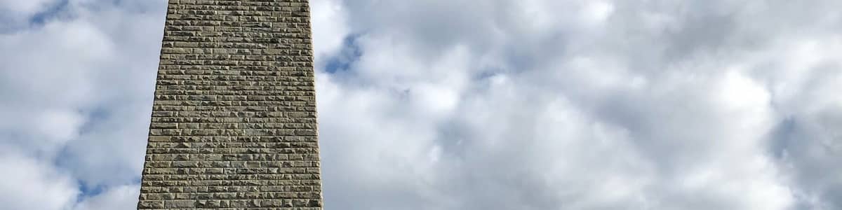 Ein hoher, steinerner Obelisk mit Wetterfahne, vor einem bewölkten Himmel.
