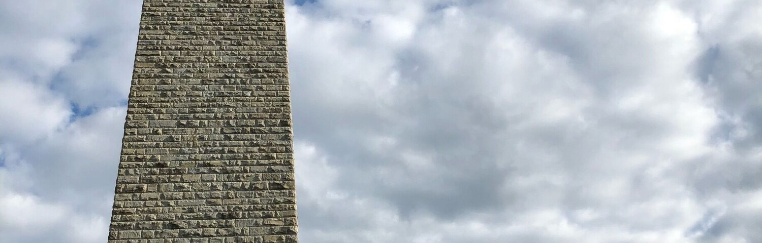 Un alto obelisco de piedra con veleta en la parte superior, sobre un cielo nublado.