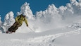 Een skiër in een gele jas en helm, die een besneeuwde helling afdaalt.