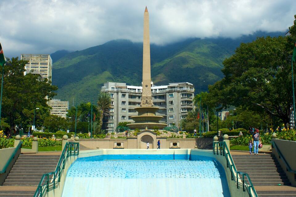 A monument with a tall obelisk in the center, surrounded by a pool and stairs, with buildings and mountains in the background.
