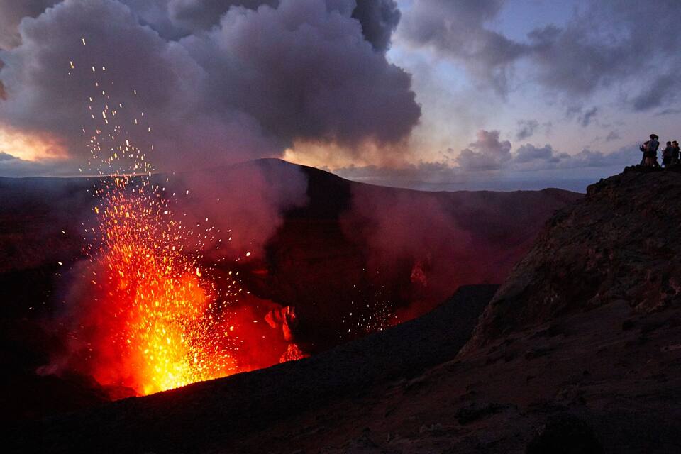 Una erupción volcánica con lava descendiendo y gente observando desde una colina.