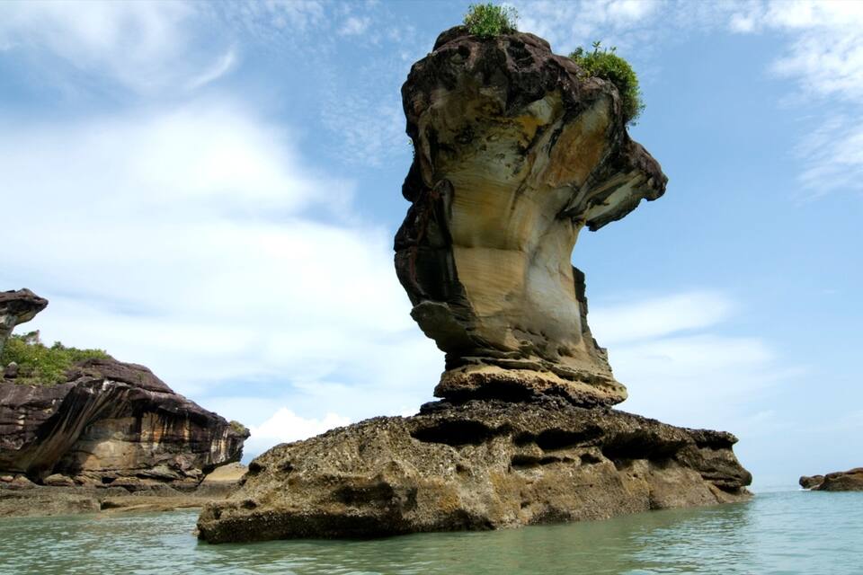 A rock formation resembling a human figure standing on a small island, surrounded by water and other rocky formations.