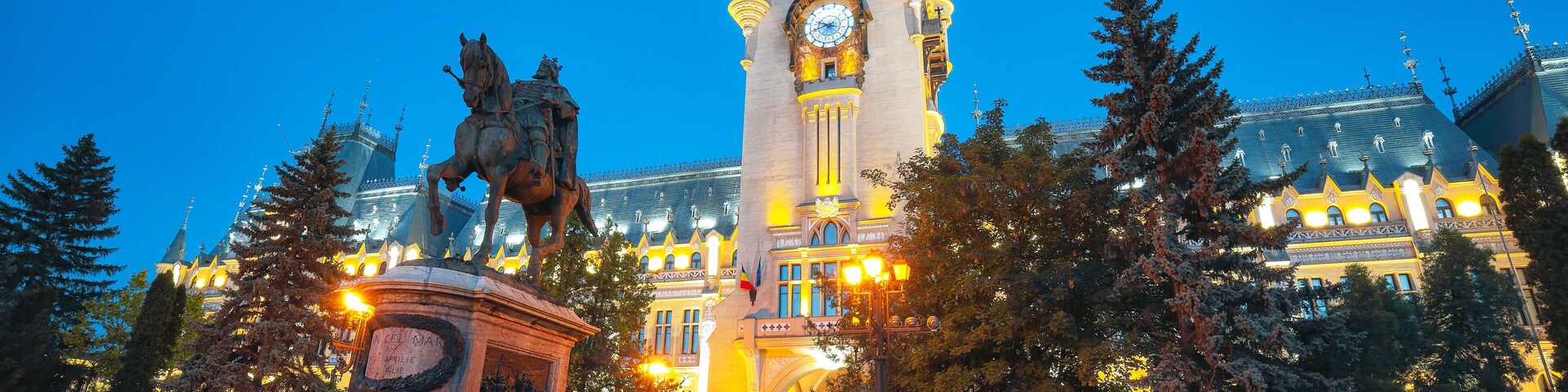 Um edifício histórico com uma torre do relógio, uma estátua de um cavalo e um parque bem cuidado com árvores.