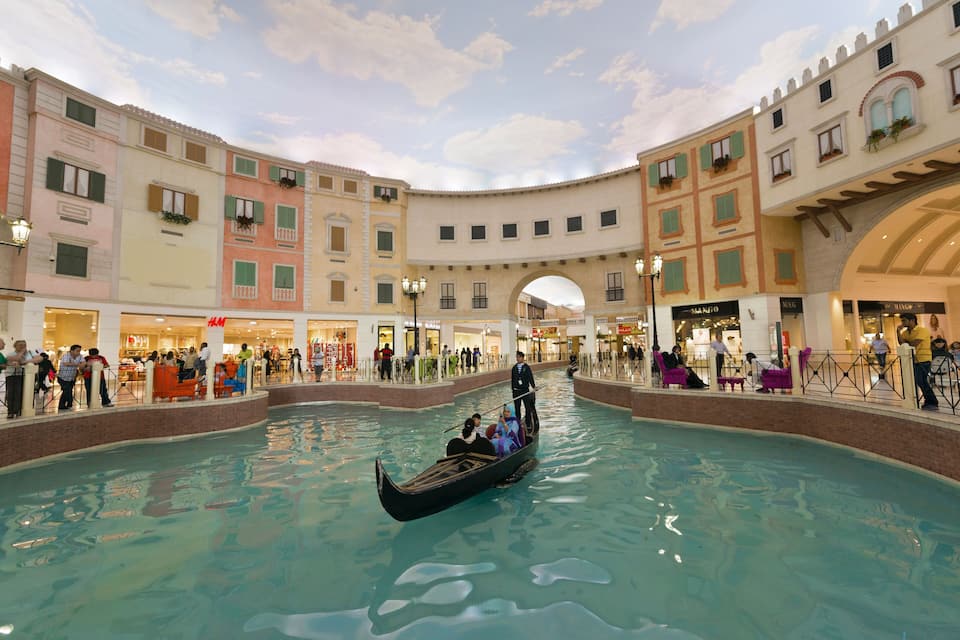 A gondola in a water-filled atrium with shoppers and buildings around.