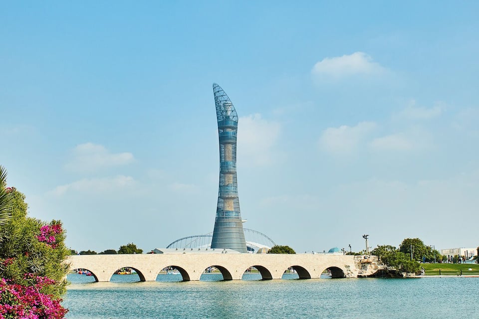A modern tower with a unique design, a bridge with arches, and a body of water with pink flowers.