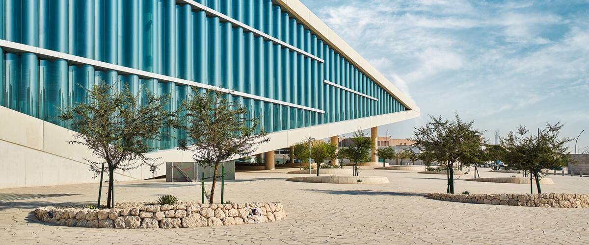 A modern building with blue vertical panels and a curved design, surrounded by a paved area with trees and stone walls.