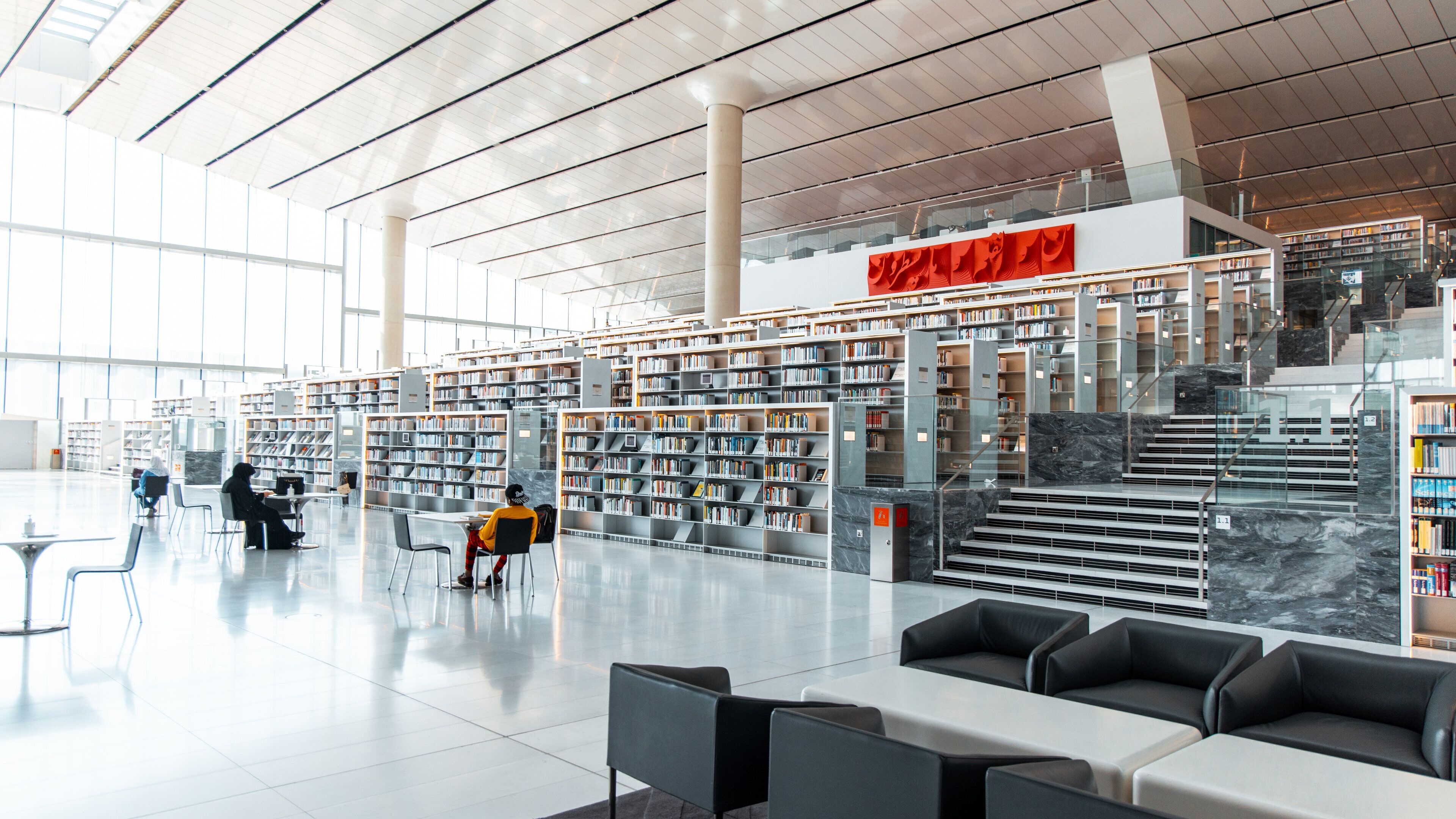 Interior de biblioteca moderna com estantes, áreas de estar e escadaria.