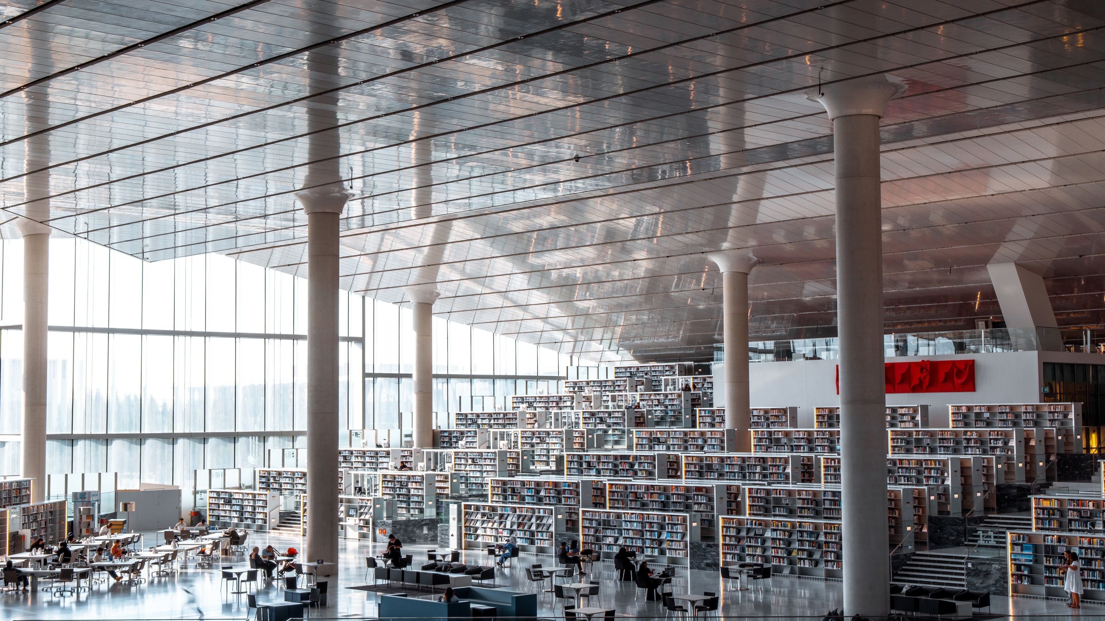 Uma biblioteca moderna com grandes janelas de vidro, vários andares de estantes e um amplo espaço para leitura.