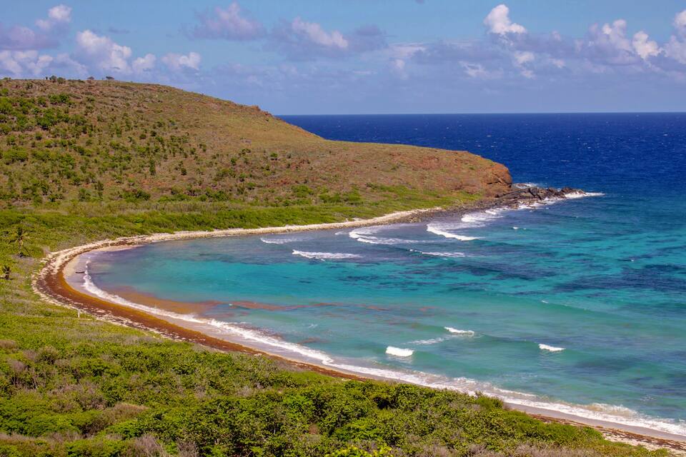 Un paisaje costero con una orilla curva, aguas turquesas cristalinas y una colina verde con vegetación escasa.