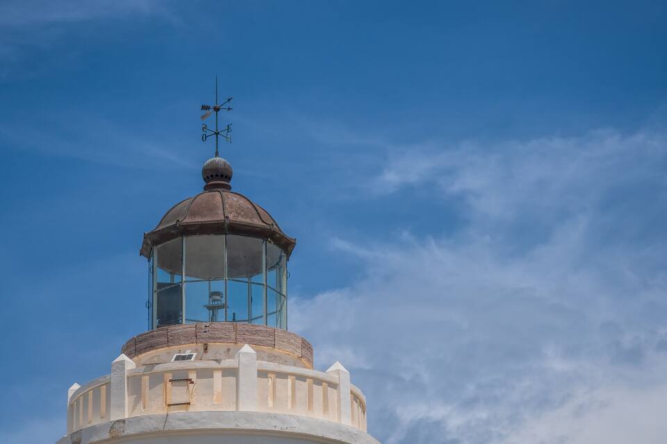 A lighthouse with a weather vane on top, set against a clear blue sky.