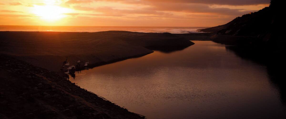 A sunset over a calm lake with a rocky shoreline and a silhouette of a person walking.