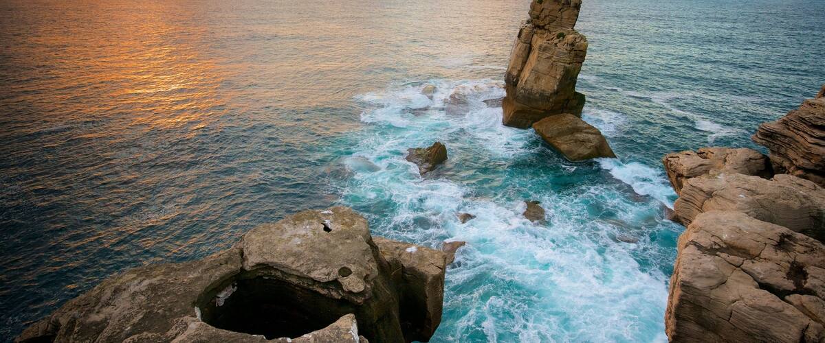 Formación rocosa costera con un agujero, olas del océano y un atardecer.