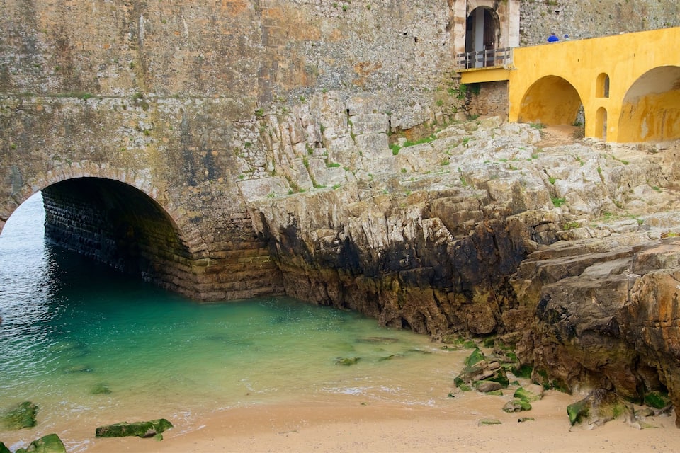Un pont de pierre enjambant un plan d’eau aux eaux claires, une plage de sable et un bâtiment jaune orné d’arcades.
