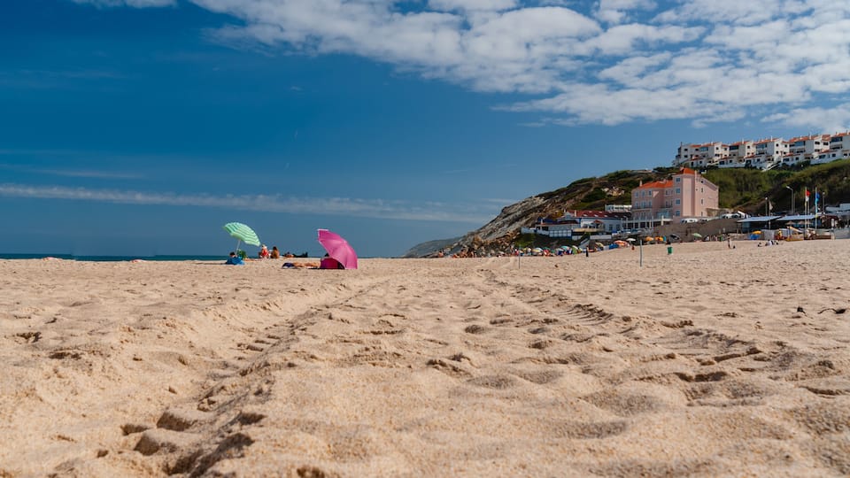 A sandy beach with footprints, a pink umbrella, and a building on a hill in the background.