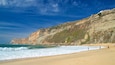 A coastal landscape with a sandy beach, rocky cliffs, and a clear blue sky.