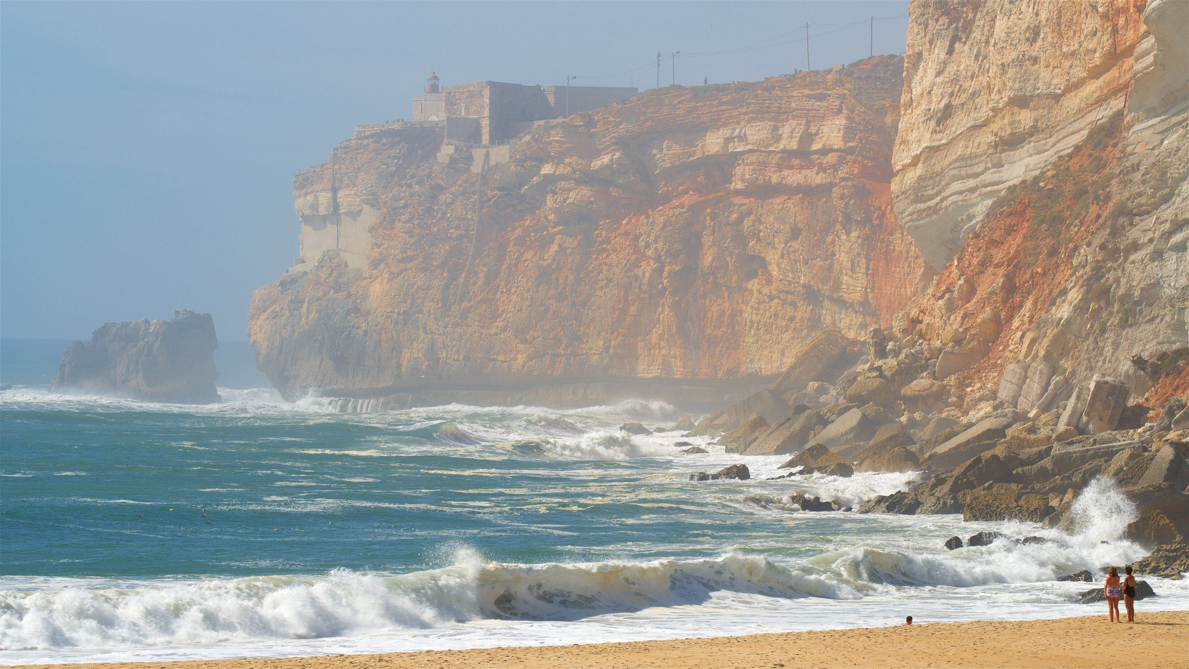 Un paysage côtier avec une falaise rocheuse, des vagues qui s’écrasent sur le rivage et un phare au sommet.