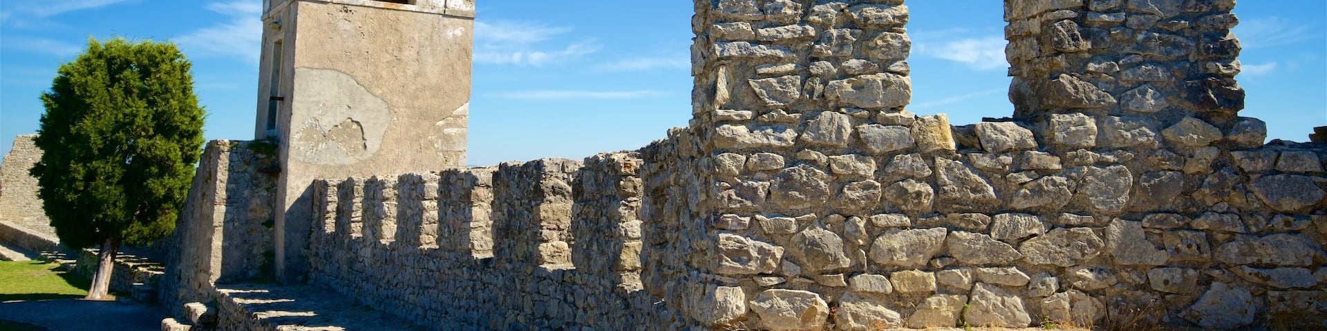A stone bell tower with a cross on top, surrounded by ruins and a clear blue sky.