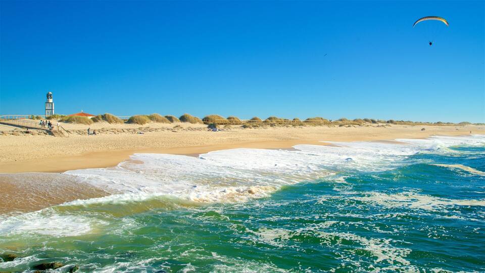 A beach with waves, a lighthouse, and a person paragliding.