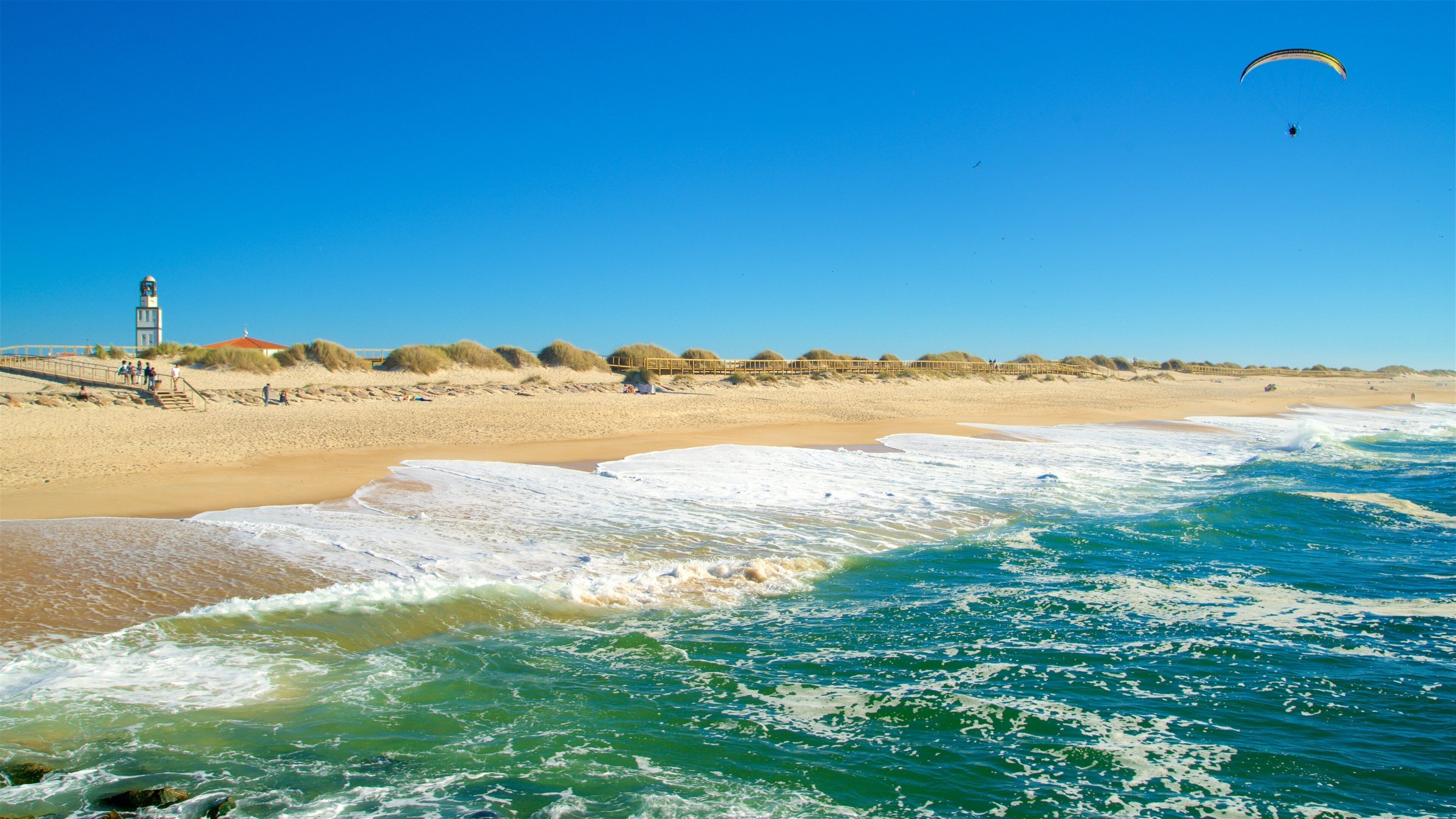 A beach with waves, a lighthouse, and a person paragliding.