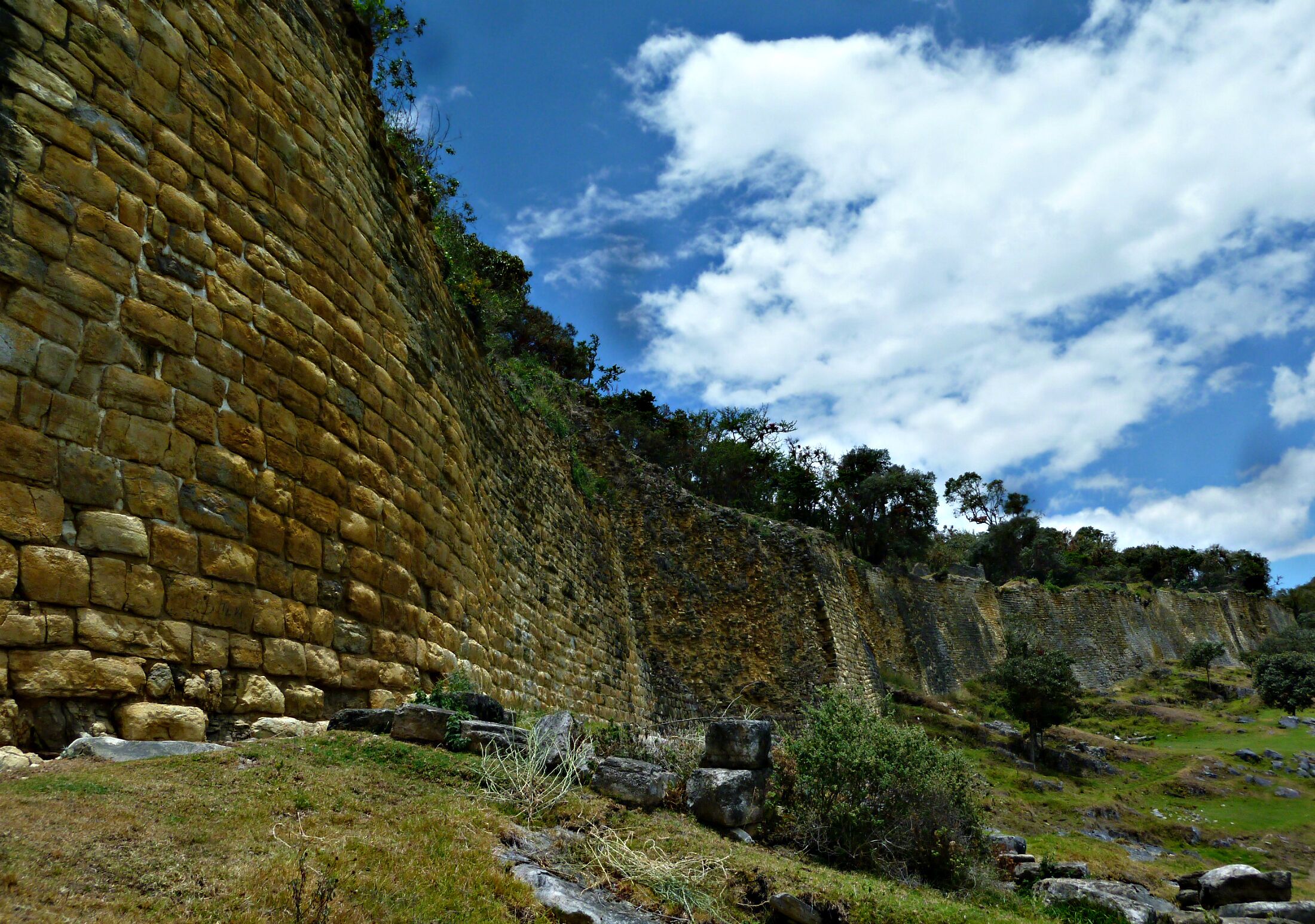 A stone wall with a grassy foreground and trees under a partly cloudy sky.