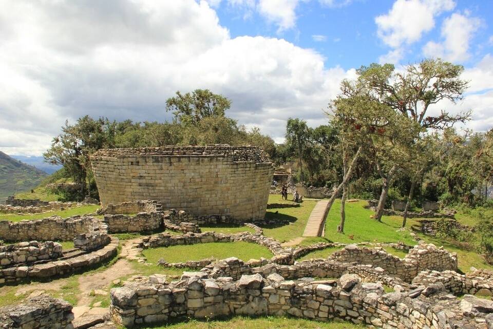 Antiguas ruinas de piedra con una gran estructura circular rodeada de edificios más pequeños de piedra, frente a un paisaje de árboles y montañas.