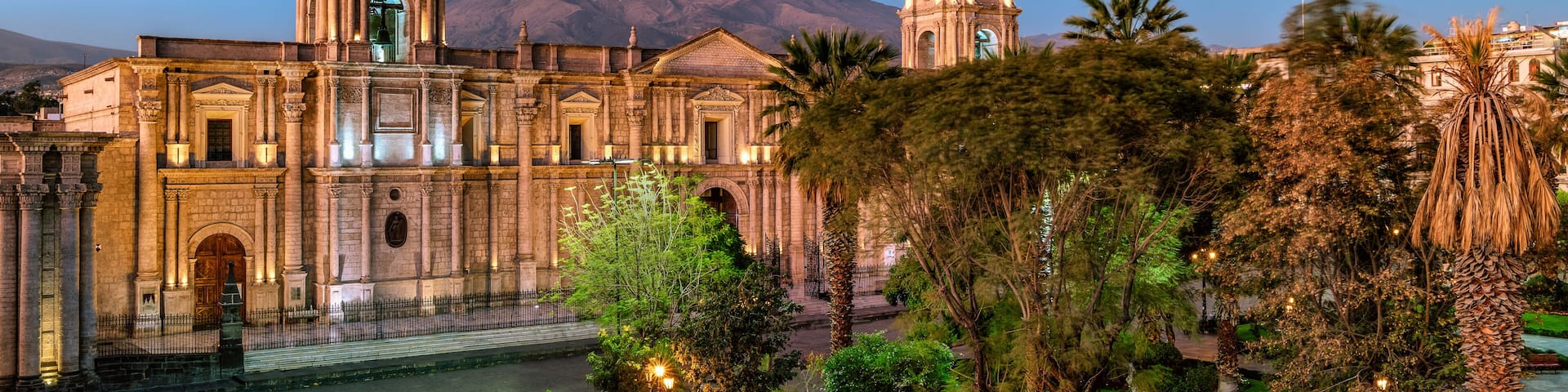A historic cathedral with two towers, a large mountain in the background, and a well-maintained garden with palm trees.