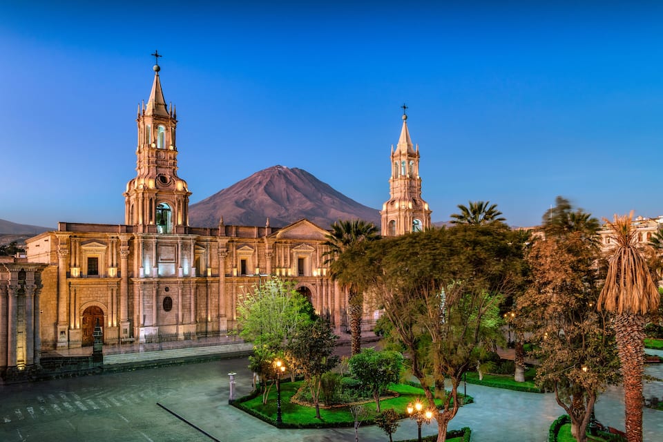 A historic cathedral with two towers, a large mountain in the background, and a well-maintained garden with palm trees.