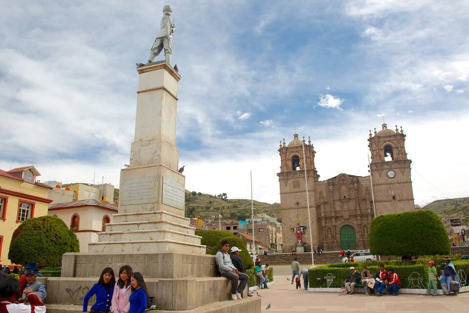 A monument with a statue, a church with two towers, and people sitting and walking around.
