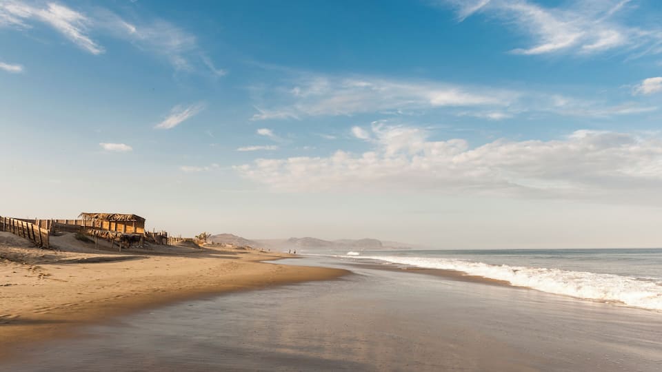 A sandy beach with a wooden structure on the left, a clear blue sky, and gentle waves.
