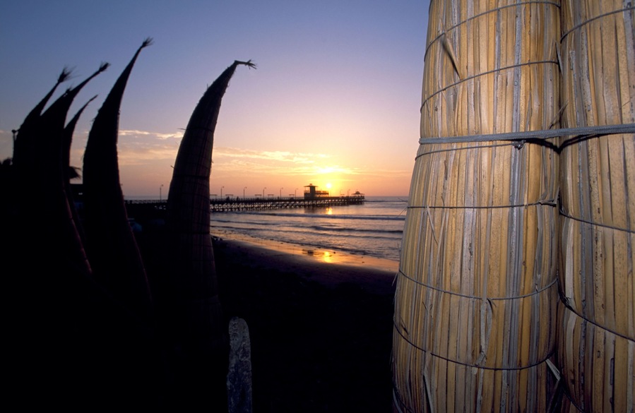 A row of traditional reed fishing boats against a sunset on the main beach front and pier of Punta Sal in Peru