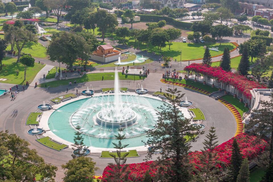 A large circular fountain surrounded by a landscaped park with trees and flower beds.