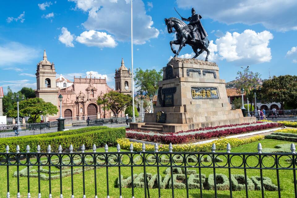 A statue of a mounted figure, a historic building, and a well-maintained garden with a fence.