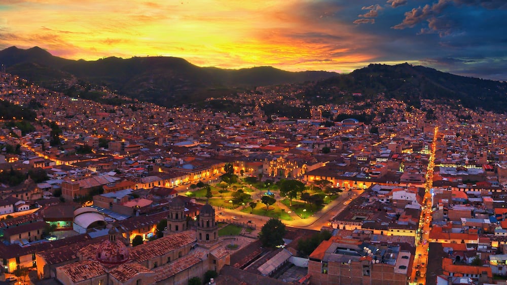 A cityscape at sunset with illuminated buildings and a mountainous backdrop.