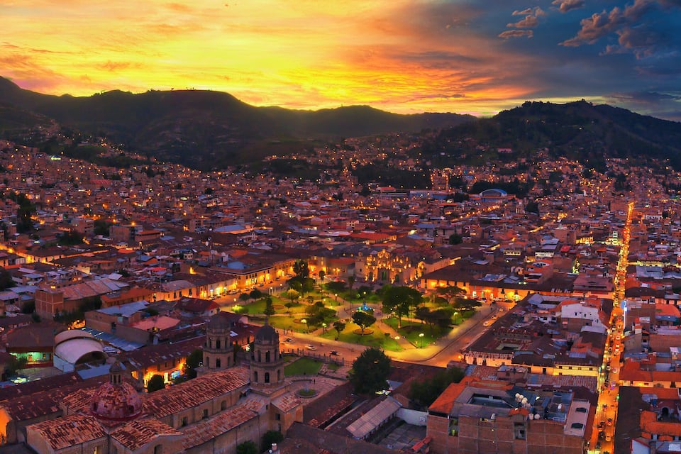 A cityscape at sunset with illuminated buildings and a mountainous backdrop.