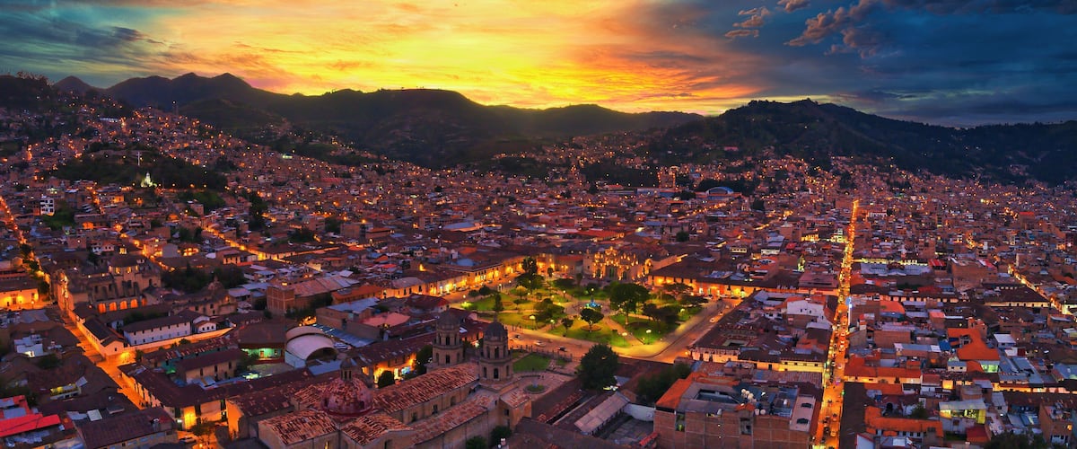 A cityscape at sunset with illuminated buildings and a mountainous backdrop.