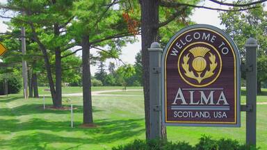 A welcome sign for Alma, Scotland, USA, surrounded by lush greenery and trees.