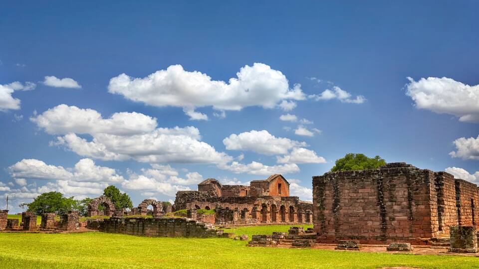 Ancient ruins with stone walls and arches, surrounded by green grass and trees under a blue sky with clouds.