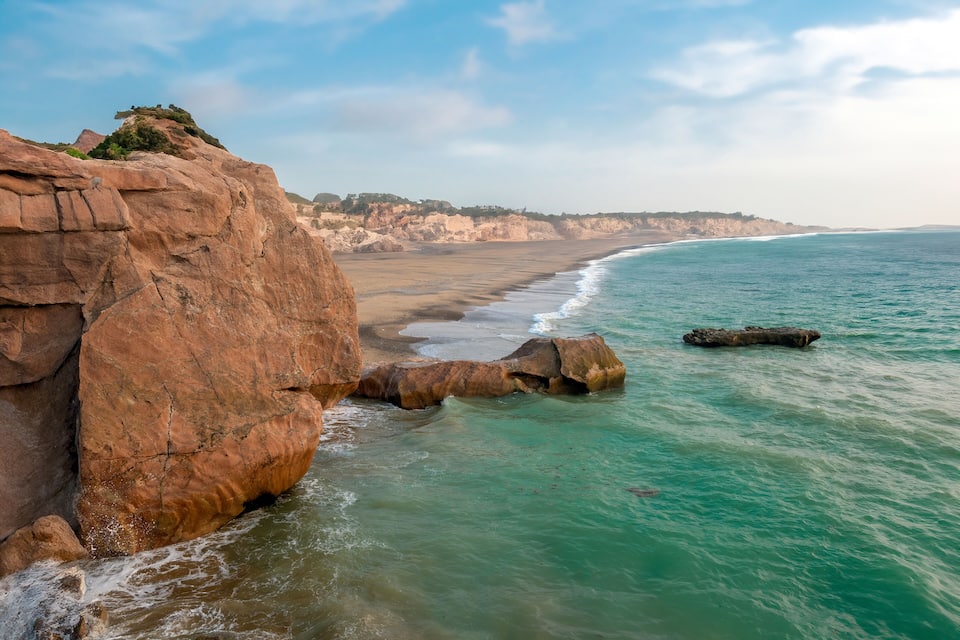 A rocky coastline with clear turquoise waters, a sandy beach, and distant cliffs.