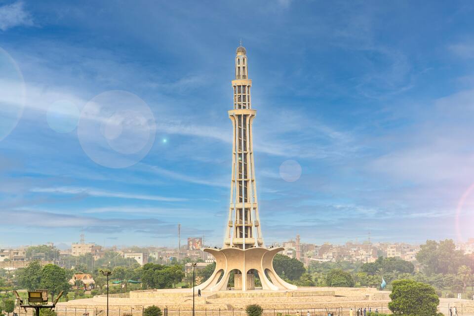 A tall tower with a dome and a spire, surrounded by a park and a cityscape in the background.