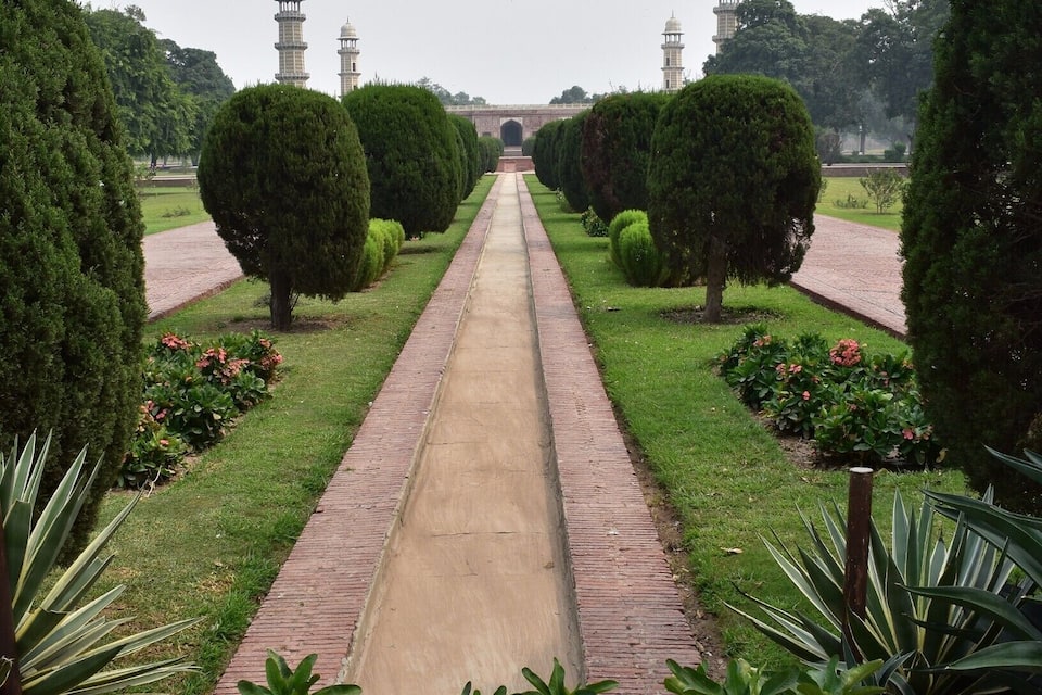 A pathway flanked by neatly trimmed hedges and potted plants, leading towards a distant structure with two minarets.