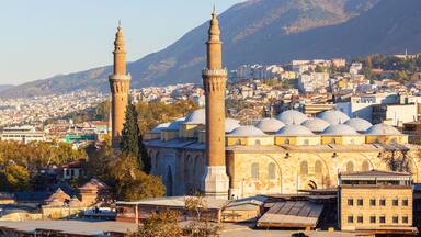 Uma mesquita com dois minaretes e várias cúpulas, rodeada por um horizonte urbano e montanhas.