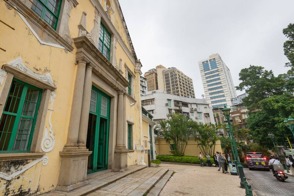 Un edifici històric groc amb portes i finestres verdes, un carrer empedrat i edificis moderns al fons.
