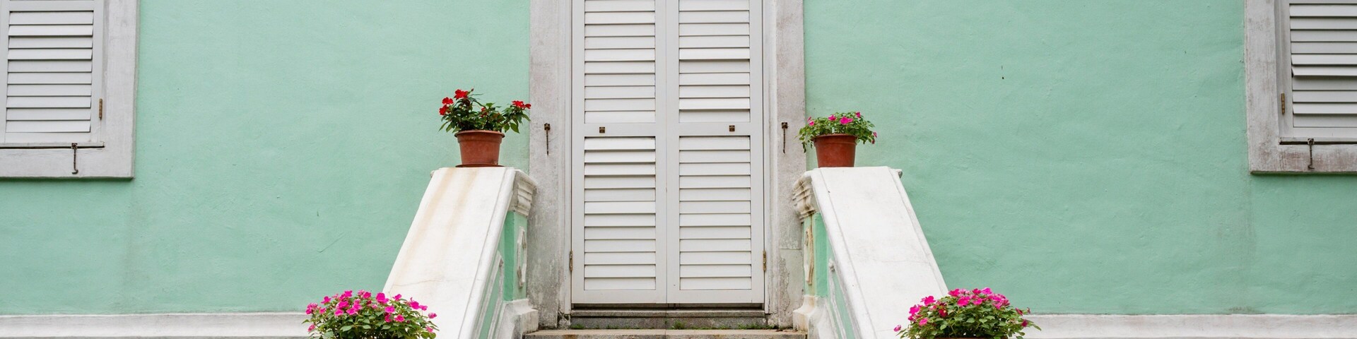 Una escalera conduce a una puerta blanca con contraventanas, flanqueada por dos macetas con flores rosadas, frente a una pared verde.