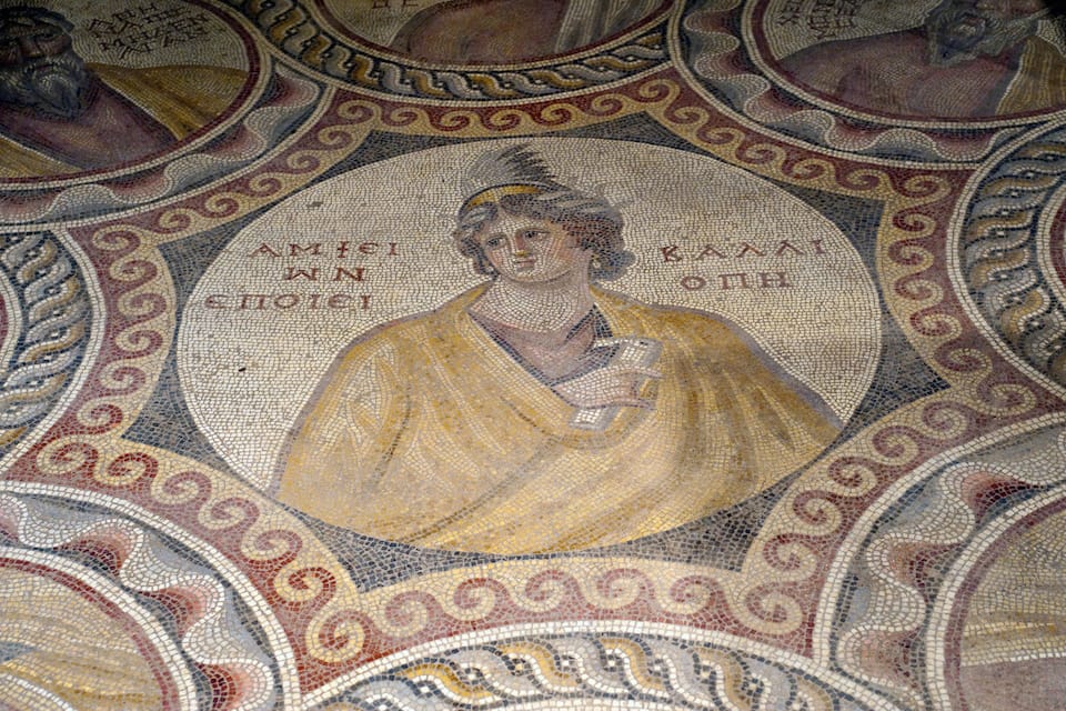 Un mosaico con una figura central sosteniendo un rollo, rodeado de diseños intrincados y otras figuras.