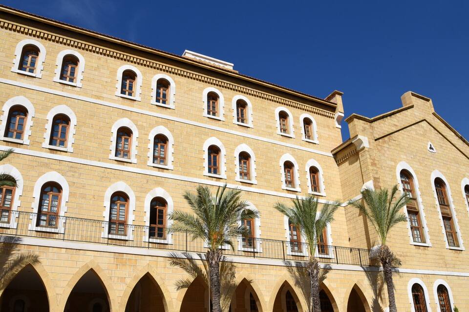 A large yellow building with multiple arched windows and balconies, surrounded by palm trees.