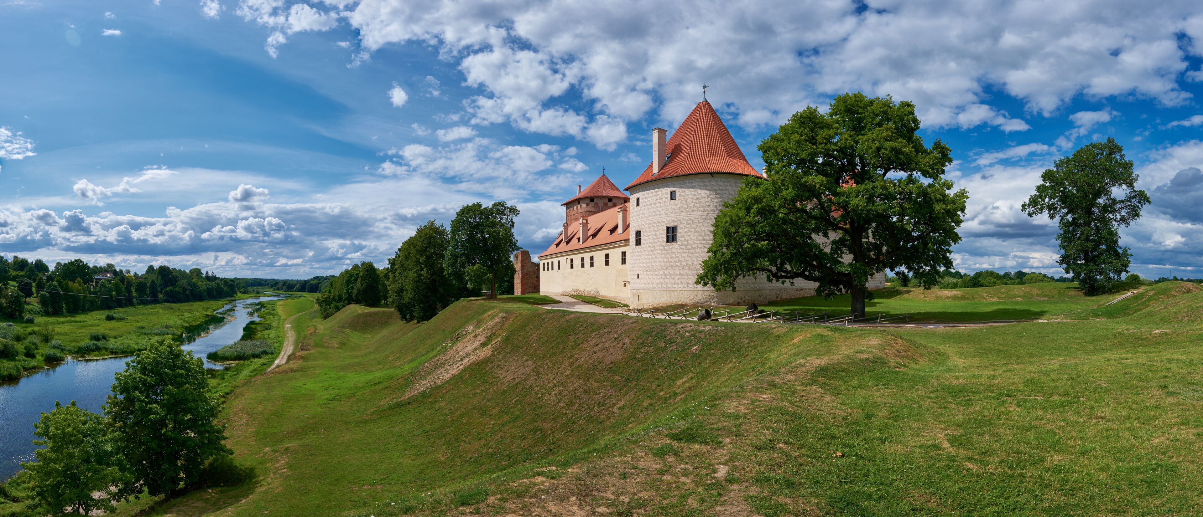 A castle with red-tiled roofs, a river, and a grassy landscape.