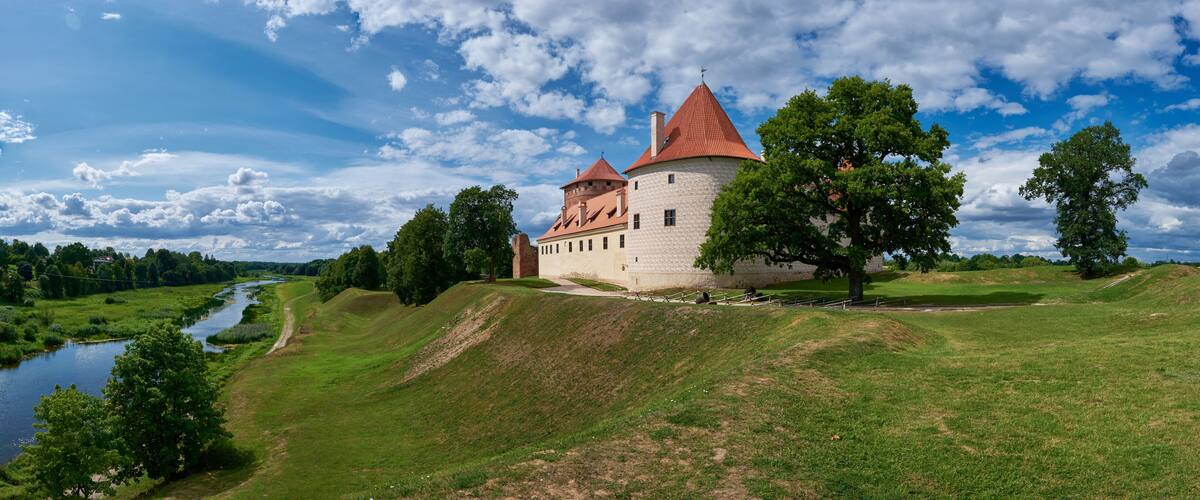 A castle with red-tiled roofs, a river, and a grassy landscape.