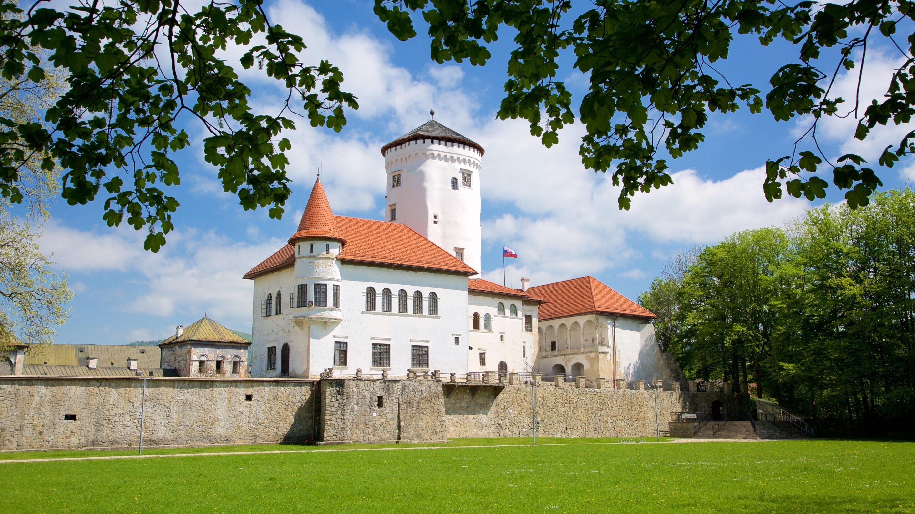 A historic castle with a central tower, surrounded by a stone wall and lush greenery.